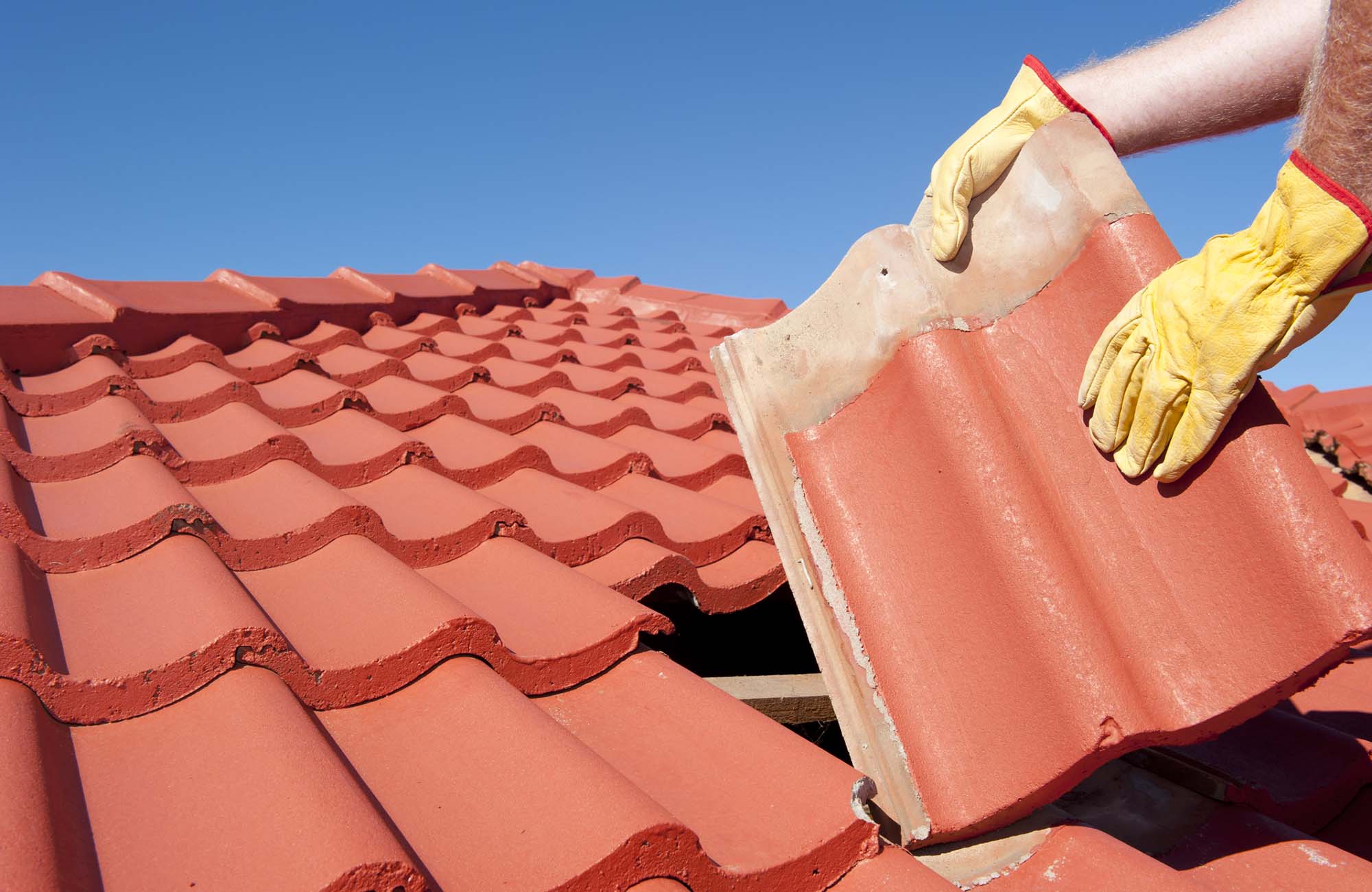 Roof repairs, worker with yellow gloves replacing red tiles or shingles on house with blue sky as background and copy space.